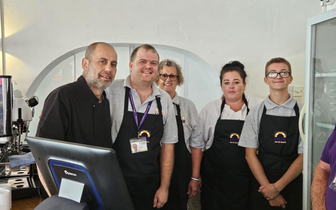 a group of staff members wearing black aprons with gold logos are posing for a picture next to a POS system at the launch of Hub on the Beach.